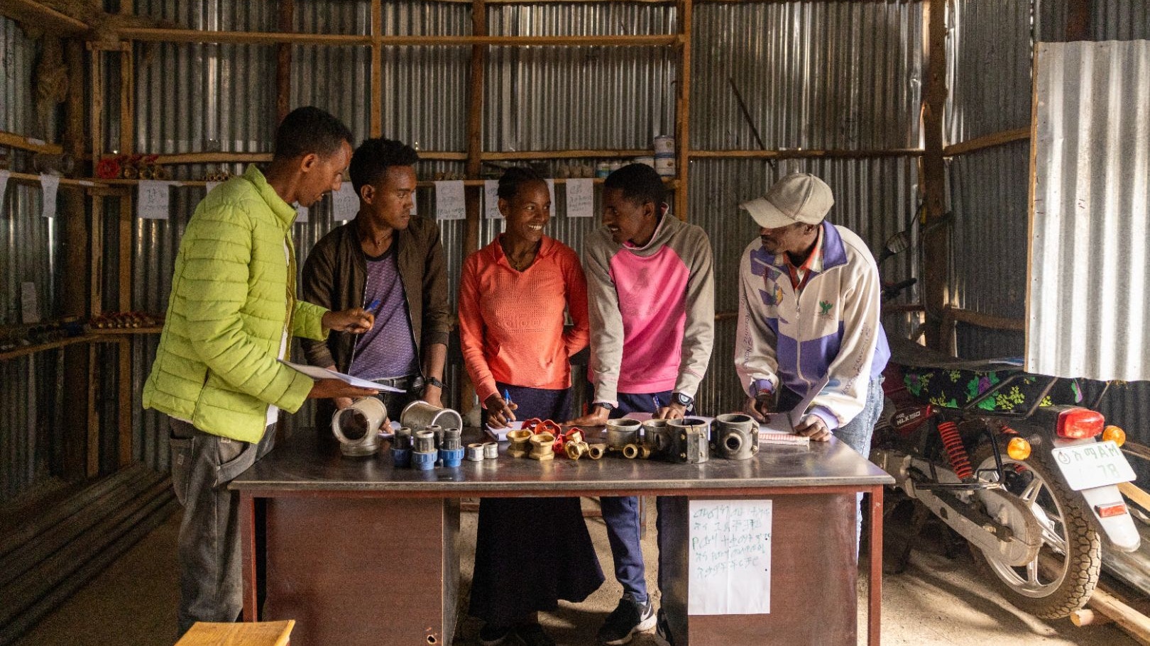 Four Ethiopian men and one woman are standing behind a table full of water pump pieces, discussing among themselves. They are members of a youth enterprise.