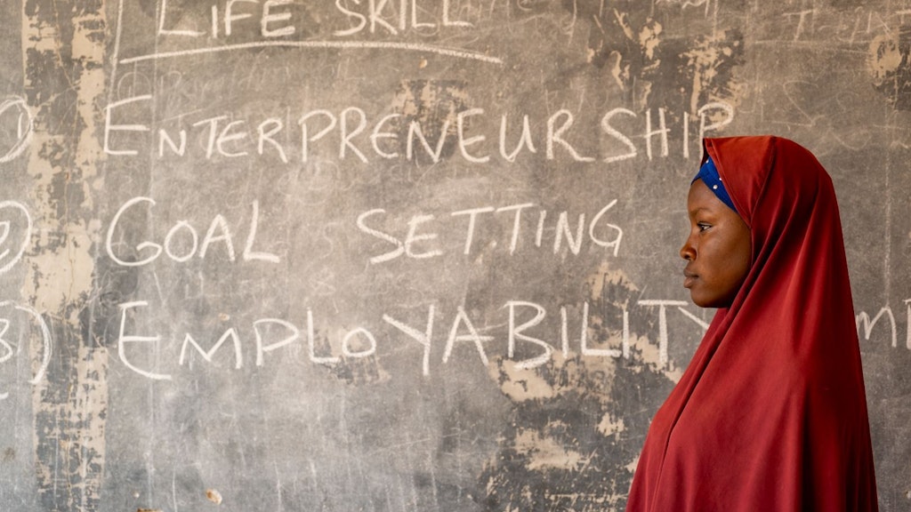 A young Nigerian woman stands in front of a blackboard, where the following is written: “Life skills: entrepreneurship, goal setting, employability”.