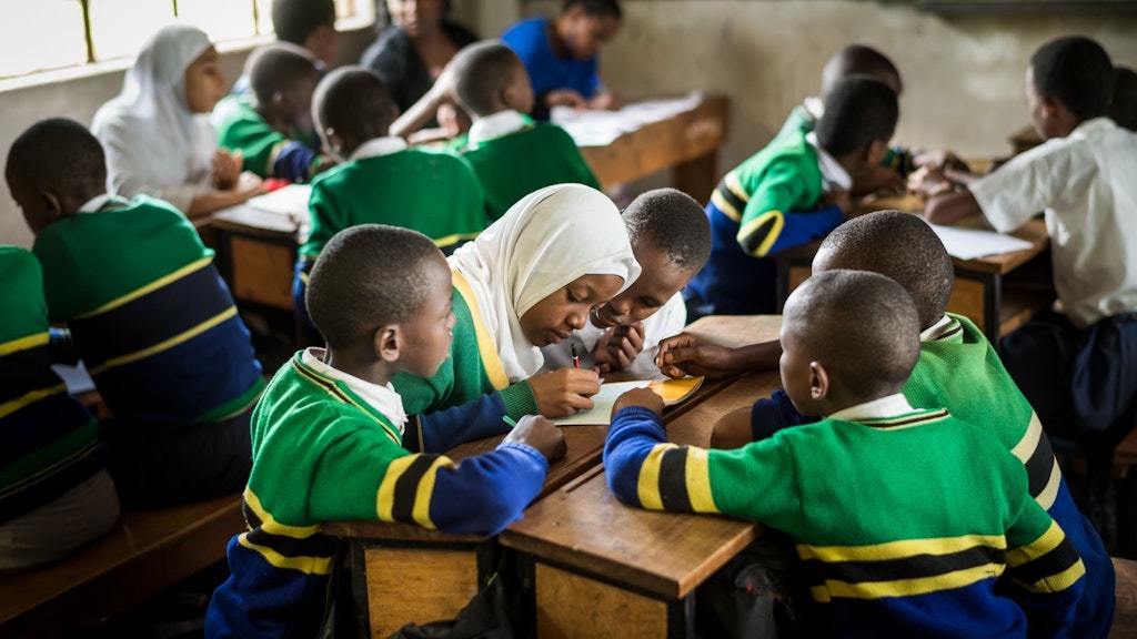 A class of primary school children sitting at their desks in small groups, in Tanzania. In one of the groups, a girl writes in a notebook and the other kids are looking at her.