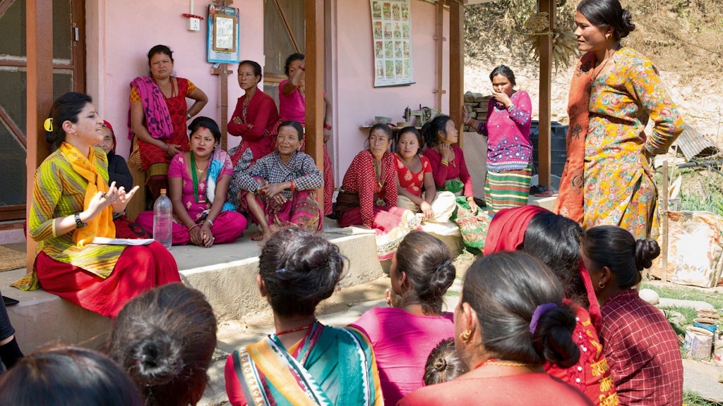Group of women in traditional Nepali dresses, sitting and standing in front of a house, listening to a woman talking about their rights.