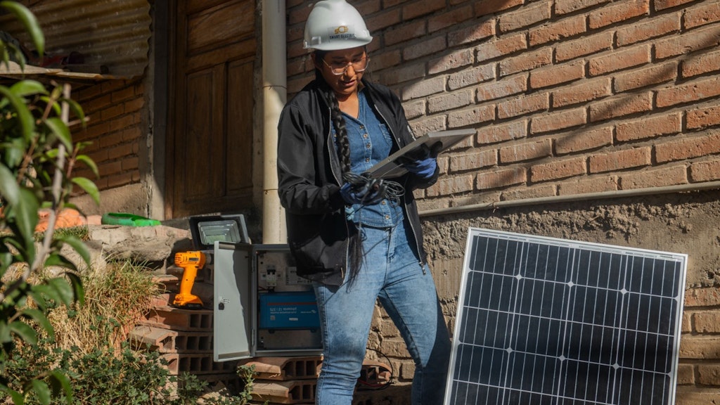 A young Bolivian woman, working as a renewable electrical systems seller, wears a helmet and glasses and holds a small solar panel. She is surrounded by another solar panel and technical equipment.