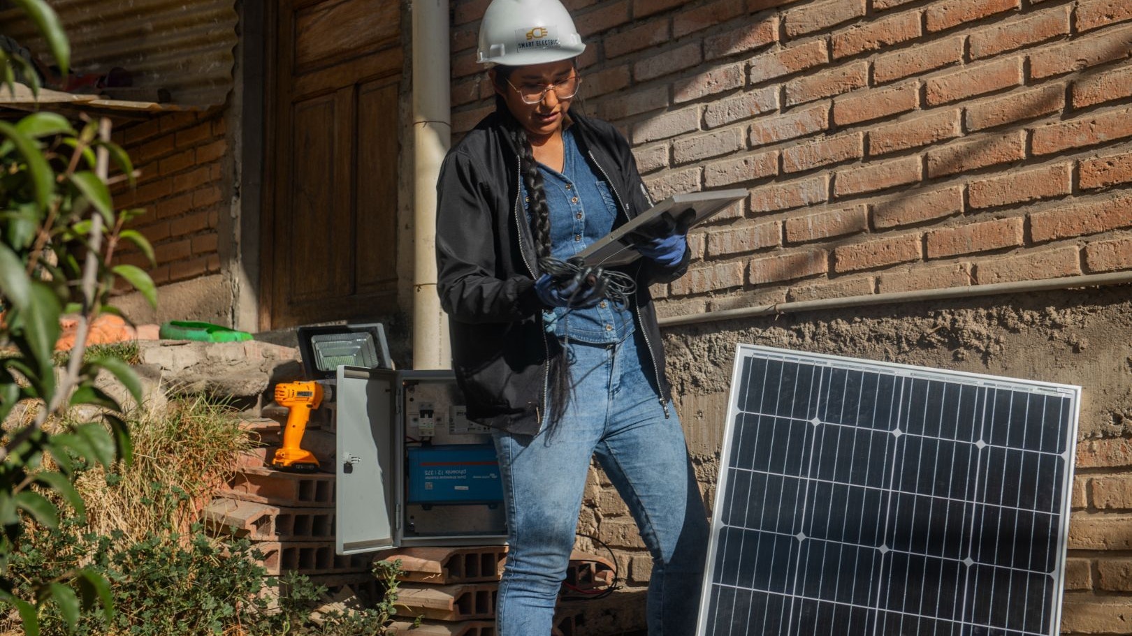 A young Bolivian woman, working as a renewable electrical systems seller, wears a helmet and glasses and holds a small solar panel. She is surrounded by another solar panel and technical equipment.