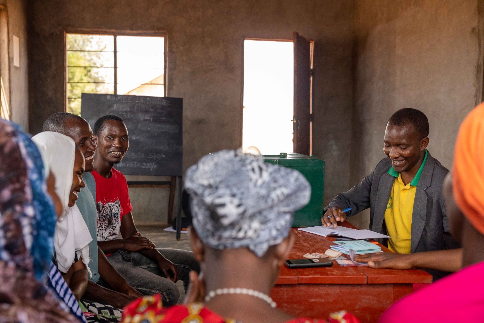 A man in a yellow and green T-Shirt is sitting at a table and counting money to distribute to smallholder farmers sitting in front of him in a village in Tanzania.
