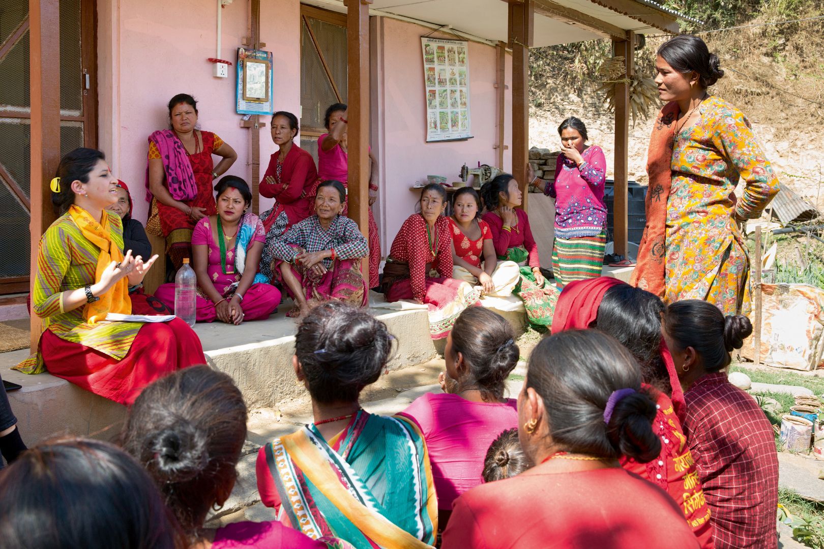Group of women in traditional Nepali dresses, sitting and standing in front of a house, listening to a woman talking about their rights.