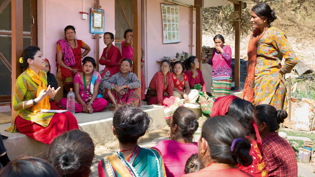 Group of women in traditional Nepali dresses, sitting and standing in front of a house, listening to a woman talking about their rights.