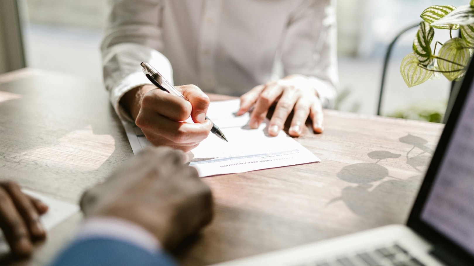 Close up of a person signing a document at a desk, in front of another person with a laptop.