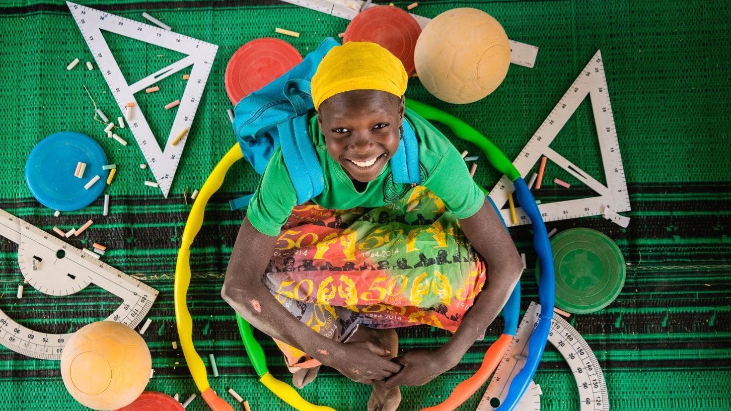 A smiley girl - seen from above – is sitting on a green carpet surrounded by various school and PE material (soccer balls, goniometers, chalks, hula hoops)
