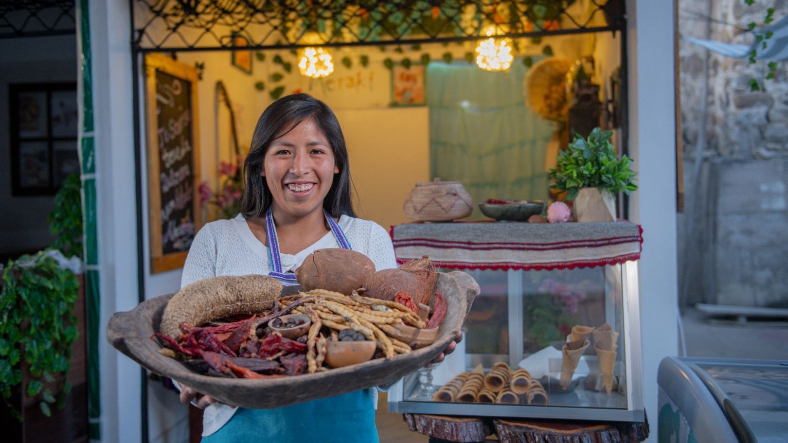 A young Bolivian woman stands smiling in front of her ice cream shop, and holds a basket with local products.