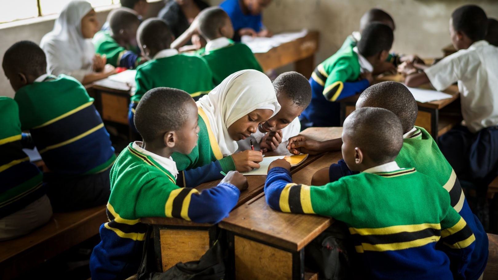A class of primary school children sitting at their desks in small groups, in Tanzania. In one of the groups, a girl writes in a notebook and the other kids are looking at her.