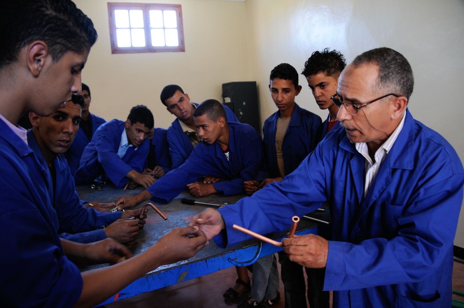 A teacher is demonstrating the handling of copper pipes to a small group of young students, wearing blue uniforms, at a vocational education and training center.